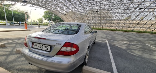 A silver car is parked in a covered parking area with a metal lattice roof. The background features trees and open spaces, and the setting appears calm and quiet.