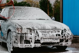 A car is covered with white foam, possibly from soap, at a car wash. The front of the car is visible, and a person is holding a red hose, rinsing the vehicle. There is a blue wall on the right side and trees in the background.