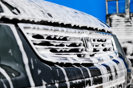 Detail of a vehicle's front grill covered in thick white soap suds. The background is a bright blue sky with part of a building visible.