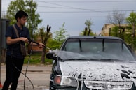 A person is using a high-pressure hose to wash a car with soap suds covering the front. The scene takes place outdoors with trees and a clear sky visible in the background.