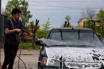 A person is using a high-pressure hose to wash a car with soap suds covering the front. The scene takes place outdoors with trees and a clear sky visible in the background.