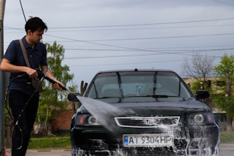 A man is using a hose to wash a black car, which is covered in soap suds. The setting appears to be outdoors, with trees and a utility pole visible in the background.