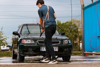 A person is washing a dark green car with a hose at an outdoor car wash. The scene includes a bright blue wall to the right and power lines in the background. Trees and buildings are visible in the distance, contributing to a suburban setting.