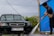 A person is washing a dark green car with a high-pressure water hose at a car wash. The car has a Ukrainian license plate and is parked on a wet concrete surface. A blue tarp partially encloses the area, and there are power lines visible in the background under a cloudy sky.