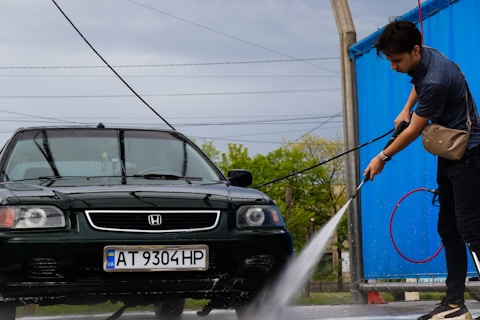 A person is washing a dark green car with a high-pressure water hose at a car wash. The car has a Ukrainian license plate and is parked on a wet concrete surface. A blue tarp partially encloses the area, and there are power lines visible in the background under a cloudy sky.
