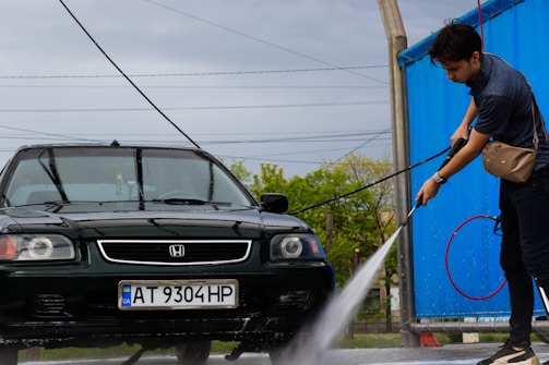 A person is washing a dark green car with a high-pressure water hose at a car wash. The car has a Ukrainian license plate and is parked on a wet concrete surface. A blue tarp partially encloses the area, and there are power lines visible in the background under a cloudy sky.