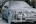 A car is covered in thick white foam, indicating it is being washed. The foam is spread over the entire surface, including the windows and wheels. The scene takes place outdoors with some greenery and buildings visible in the background.