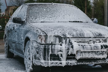 A car is covered in thick white foam, indicating it is being washed. The foam is spread over the entire surface, including the windows and wheels. The scene takes place outdoors with some greenery and buildings visible in the background.