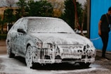 A car is covered in soap suds, presumably being washed. Beside the car, a person in casual clothing is holding a water hose. The background shows some trees and a blue wall or curtain.