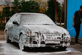 A car is covered in soap suds, presumably being washed. Beside the car, a person in casual clothing is holding a water hose. The background shows some trees and a blue wall or curtain.