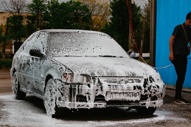 A car is covered in soap suds, presumably being washed. Beside the car, a person in casual clothing is holding a water hose. The background shows some trees and a blue wall or curtain.