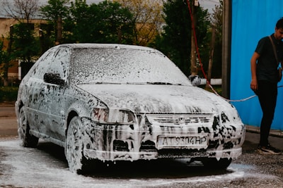 A car is covered in soap suds, presumably being washed. Beside the car, a person in casual clothing is holding a water hose. The background shows some trees and a blue wall or curtain.