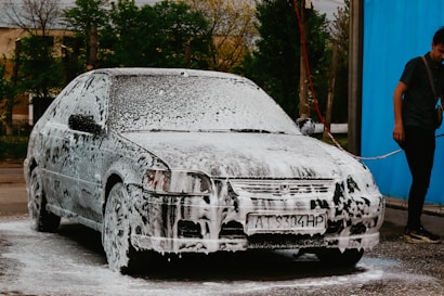 A car is covered in soap suds, presumably being washed. Beside the car, a person in casual clothing is holding a water hose. The background shows some trees and a blue wall or curtain.