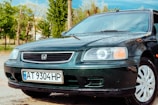 A dark green Honda car is parked on a wet pavement, with water droplets visible on its surface. The car features white alloy wheels and a Ukrainian license plate under the front grille. In the background, there are green trees and a partially visible building, suggesting an outdoor setting.
