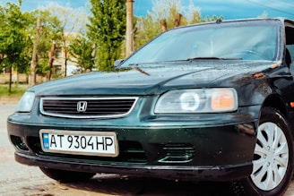 A dark green Honda car is parked on a wet pavement, with water droplets visible on its surface. The car features white alloy wheels and a Ukrainian license plate under the front grille. In the background, there are green trees and a partially visible building, suggesting an outdoor setting.