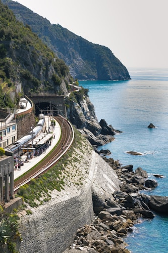 A scenic coastal train track runs along a steep rocky cliff with lush greenery. The track curves gently, passing through a tunnel and a small train station filled with several people. The clear blue sea stretches out to the horizon on the right.