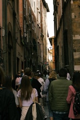 A narrow, crowded alleyway flanked by tall, old brick buildings. A group of people, including tourists with backpacks and jackets, walk down the cobblestone path, creating a lively atmosphere.