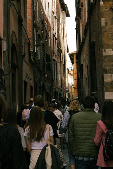 A narrow, crowded alleyway flanked by tall, old brick buildings. A group of people, including tourists with backpacks and jackets, walk down the cobblestone path, creating a lively atmosphere.