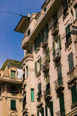 A shot of a classic European-style apartment building with ornate architectural details. The facade features several windows with green shutters and small balconies with intricate metal railings. The building is bathed in sunlight, casting slight shadows and highlighting the texture of the walls. Wires hang across the view and a clear blue sky forms the backdrop.