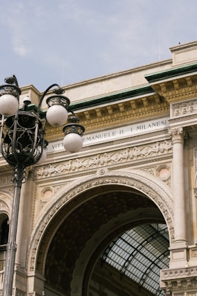 An ornate architectural archway features intricate designs and sculptures, partly visible are the words 'Vittorio Emanuele II'. In the foreground, a vintage-style street lamp with multiple spherical lights complements the classic aesthetic.