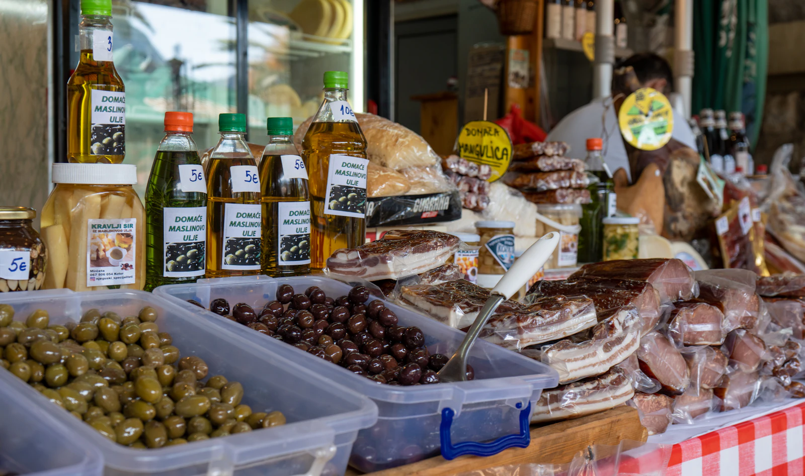 Market stall display of olives, cured meats, cheese, and bottled olive oil