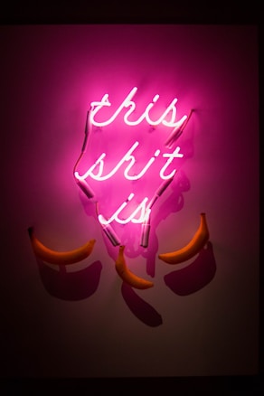 A neon light sign emits a bright pink glow with the words in cursive. Below the neon sign, several bananas are strategically placed to create an artistic and humorous display with shadows cast on the wall.
