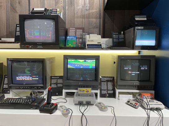 A collection of vintage computer monitors and gaming consoles is set up on display. The monitors show various types of retro graphics and games. There is a noticeable presence of controls, such as joysticks and gaming pads, with visible connecting cables, reflecting the technology from a past era.