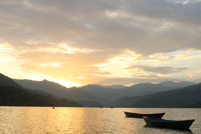 A serene lake scene at sunrise, featuring calm waters with a gentle reflection of the sky. There are small boats floating on the water, and mountains in the background are silhouetted against the brightening sky.