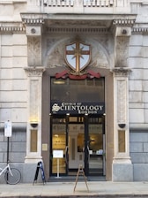 The entrance of a building with ornate architectural features, including decorative columns and a shield emblem above the door. The signage reads 'Church of Scientology London,' and there are glass doors allowing a glimpse of the interior. A bicycle is parked outside, and there are signs related to building access.