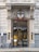 The entrance of a building with ornate architectural features, including decorative columns and a shield emblem above the door. The signage reads 'Church of Scientology London,' and there are glass doors allowing a glimpse of the interior. A bicycle is parked outside, and there are signs related to building access.