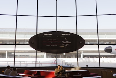 A modern airport departure lounge with large floor-to-ceiling windows showcasing an outdoor view of the airfield. A vintage-style departure board with arrivals and departures information is prominently displayed. Inside the lounge, several people can be seen sitting and relaxing on red cushioned seating. The nose of an airplane is visible outside, suggesting a busy airport environment.