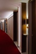 A hotel corridor with a modern design features a long red carpet and minimalist wooden panels. The lighting is soft, casting a warm glow along the hallway. Room numbers are displayed on the wood panels.