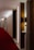 A hotel corridor with a modern design features a long red carpet and minimalist wooden panels. The lighting is soft, casting a warm glow along the hallway. Room numbers are displayed on the wood panels.