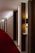 A hotel corridor with a modern design features a long red carpet and minimalist wooden panels. The lighting is soft, casting a warm glow along the hallway. Room numbers are displayed on the wood panels.