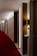 A hotel corridor with a modern design features a long red carpet and minimalist wooden panels. The lighting is soft, casting a warm glow along the hallway. Room numbers are displayed on the wood panels.