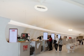 A spacious food hall with a curved modern ceiling featuring several counter service food stalls. There are digital menu screens above each counter. People are standing at the counters, some ordering food. There are retro-style white chairs and small tables in the seating area.