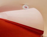 A minimalist interior space featuring a curved red carpeted walkway with an overhead sign displaying 'TWA'. The walls and ceiling are smooth and pale, creating a futuristic and clean appearance.