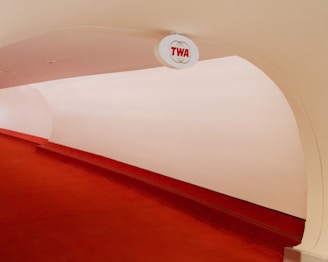 A minimalist interior space featuring a curved red carpeted walkway with an overhead sign displaying 'TWA'. The walls and ceiling are smooth and pale, creating a futuristic and clean appearance.
