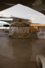 A translucent glass door with the TWA logo is visible, reflecting an airport tarmac in the background. The logo features large letters and a globe design, along with the word 'HOTEL' below it. The image captures a nostalgic atmosphere with the reflection suggesting a busy airport scene.