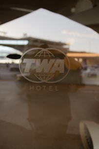 A translucent glass door with the TWA logo is visible, reflecting an airport tarmac in the background. The logo features large letters and a globe design, along with the word 'HOTEL' below it. The image captures a nostalgic atmosphere with the reflection suggesting a busy airport scene.