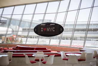 A modern airport terminal interior with large, expansive windows offering a view of the tarmac and parts of the airport buildings. The space features sleek, white and red furniture, including tables and chairs, arranged in a stylish, contemporary manner. An oval-shaped digital departure and arrival board is prominently displayed, providing flight information. The environment appears calm and orderly.