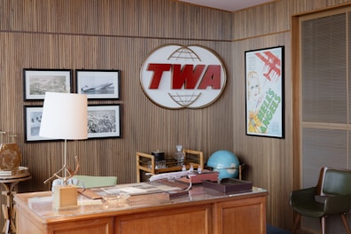 A vintage-style office setting with a wooden desk and chair, accompanied by a retro-style globe. A large TWA logo is mounted on the wood-paneled wall, surrounded by black-and-white photographs of airplanes. A poster with red airplane graphics and text is framed on the wall. A white table lamp sits on the desk along with other office accessories.