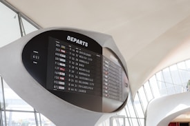 A large, modern airport departure board displays flight information, including departure times, flight numbers, and destinations. The board is set against a sleek, curved architectural background with large windows allowing natural light to illuminate the area.