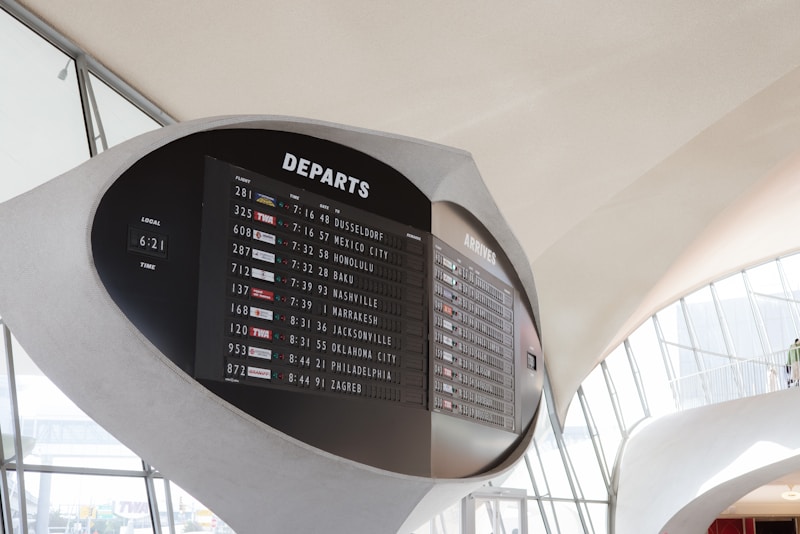 A large, modern airport departure board displays flight information, including departure times, flight numbers, and destinations. The board is set against a sleek, curved architectural background with large windows allowing natural light to illuminate the area.