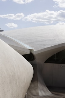 A modern architectural structure featuring smooth, flowing curves and large concrete surfaces under a partly cloudy sky. The design emphasizes sleek, aerodynamic forms with a focus on minimalism and elegance.