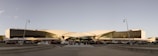 A futuristic, symmetrically designed airport terminal with a unique, curvy roof structure and expansive glass facade. Several cars are parked in front of the terminal, and a wide pathway leads to the entrance. Minimalist street lamps line the foreground, and the sky is clear, suggesting good weather.
