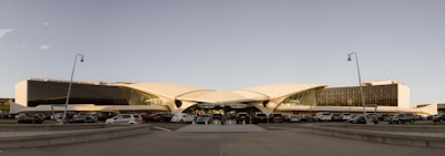 A futuristic, symmetrically designed airport terminal with a unique, curvy roof structure and expansive glass facade. Several cars are parked in front of the terminal, and a wide pathway leads to the entrance. Minimalist street lamps line the foreground, and the sky is clear, suggesting good weather.