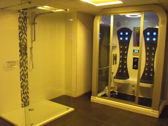 A modern bathroom features a glass-enclosed shower on the left with a sleek design and decorative tiles. On the right, there is a high-tech steam shower cabin equipped with multiple control panels and LED lights.