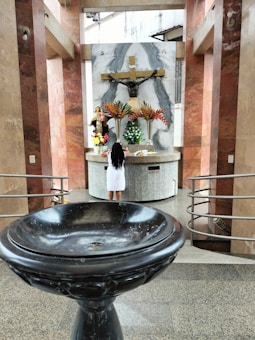 A person stands in front of a religious altar adorned with a large crucifix and multiple floral arrangements. The altar area is framed with stone columns and a decorative basin is prominently displayed in the foreground.