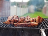 A barbecue grill with several pieces of meat being cooked, surrounded by a smoky atmosphere. The background shows a blurred outdoor setting with grass and a fence.